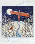 Festive Christmas card by Joanne Wishart featuring a snowy path leading to the Angel of the North, with a curious robin observing. The landmark, bathed in bright moonlight, stands tall over snow covered Gateshead hill.