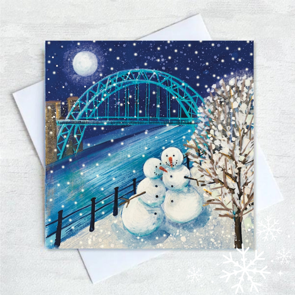 Two snowmen stand smiling, in the snow, at the edge of the River Tyne. The moon is full and shines above the Tyne Bridge and a snow covered tree in the foreground. 