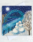 Two snowmen stand smiling, in the snow, at the edge of the River Tyne. The moon is full and shines above the Tyne Bridge and a snow covered tree in the foreground. 