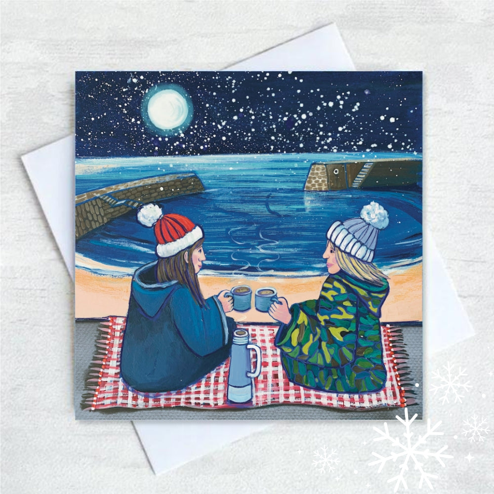 Two figures, one with long brown hair and the other with blond hair and woolly hats sit on a rug in the foreground with steaming mugs held close together and a thermos flask between them. The full moon lights up a sky of snowflakes and the beach of Cullercoats Bay and the two piers.