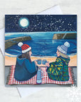 Two figures, one with long brown hair and the other with blond hair and woolly hats sit on a rug in the foreground with steaming mugs held close together and a thermos flask between them. The full moon lights up a sky of snowflakes and the beach of Cullercoats Bay and the two piers.