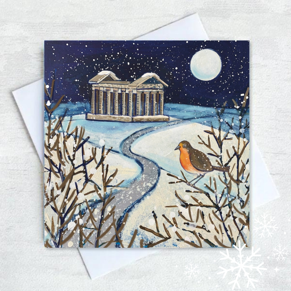 A path leads through the snow past Penshaw Monument on the horizon. In the foreground a robin perches on the bare branches of a bush and the full moon illuminates the snowflakes in the dark night sky.