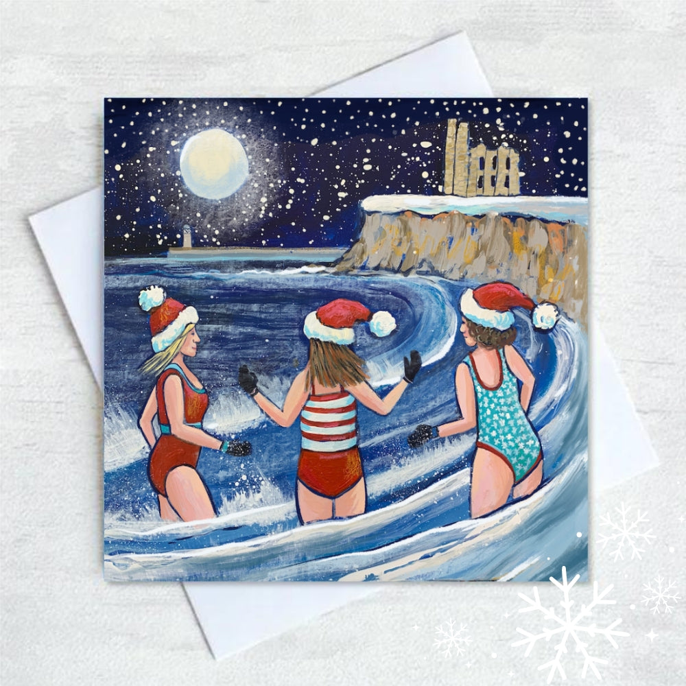 A trio enjoy a festive swim in the waves at King Edward's Bay in Tynemouth. The card features Tynemouth priory in the distance on a cliff and a full moon over the lighthouse. Snow is falling in the dark night sky.