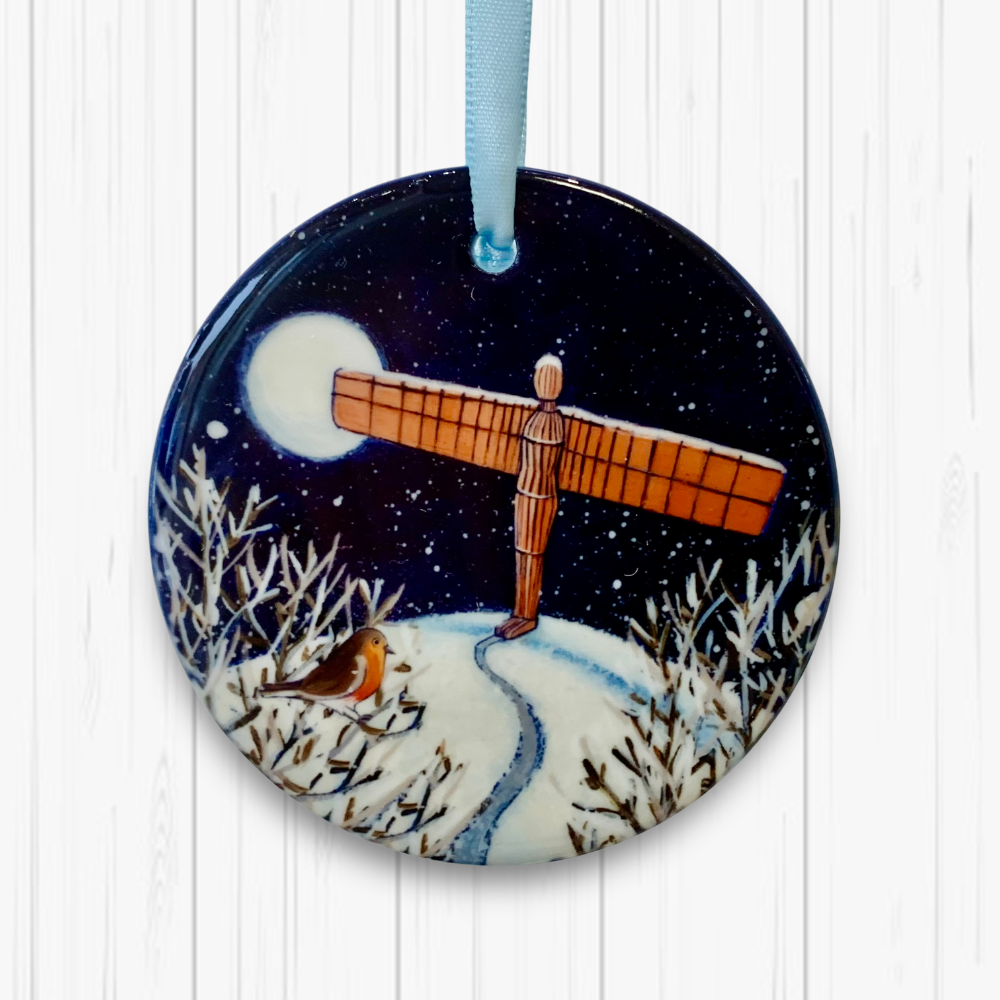 Circular ceramic decoration against a white background. It features the Angel of the North on a snowy hill with a robin nearby. There is a full moon in the dark and snowy sky. 