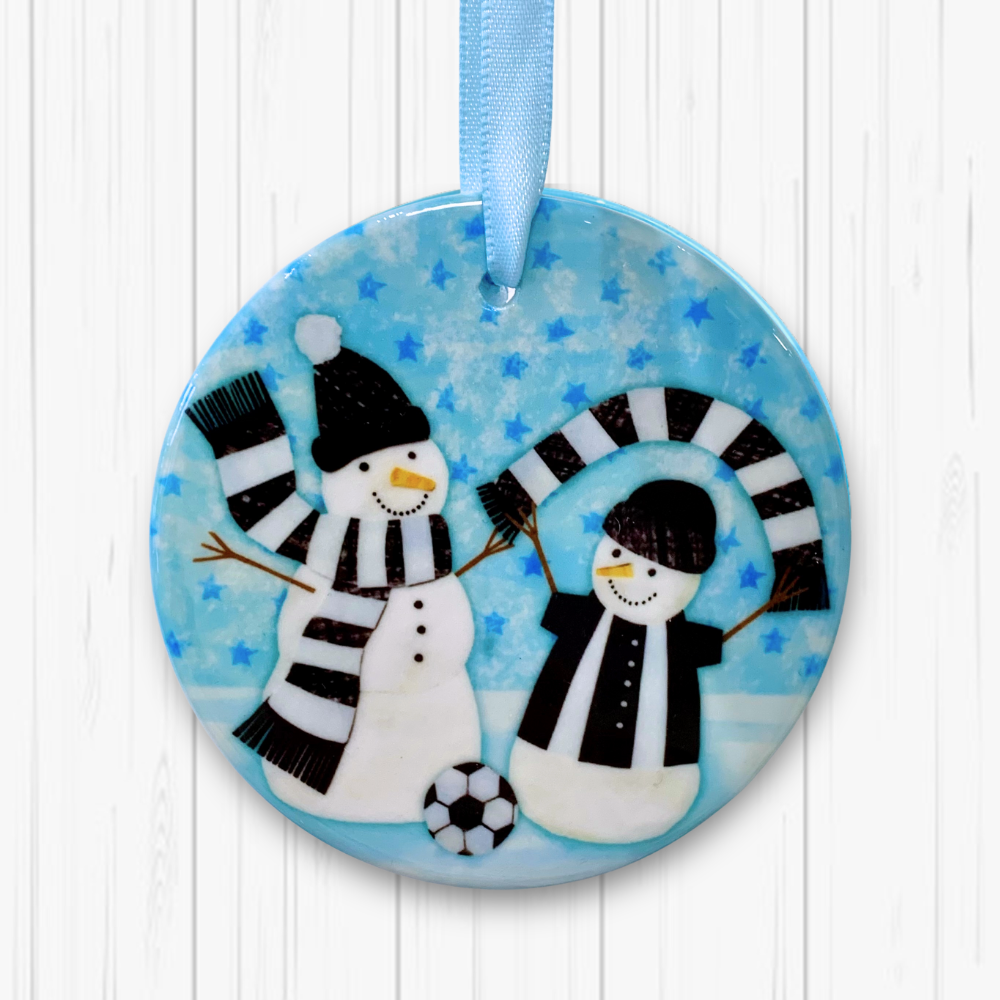 Circular ceramic decoration hung against a white background. Happy snowmen are wearing black and white scarves and football kit with a football at their feet. 