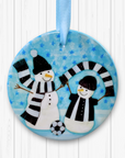 Circular ceramic decoration hung against a white background. Happy snowmen are wearing black and white scarves and football kit with a football at their feet. 