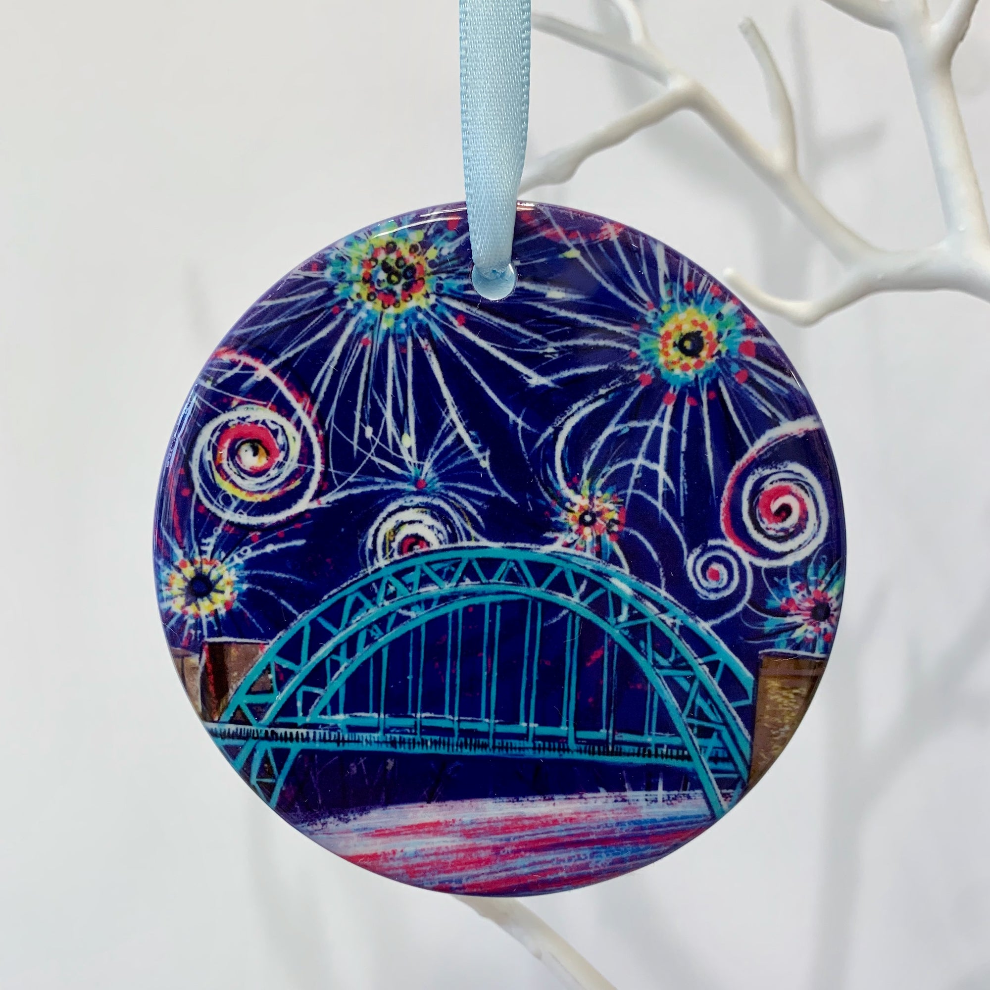 A circular ceramic decoration hung on a white branch. It features the Tyne Bridge under a dark blue sky of colourful fireworks. 