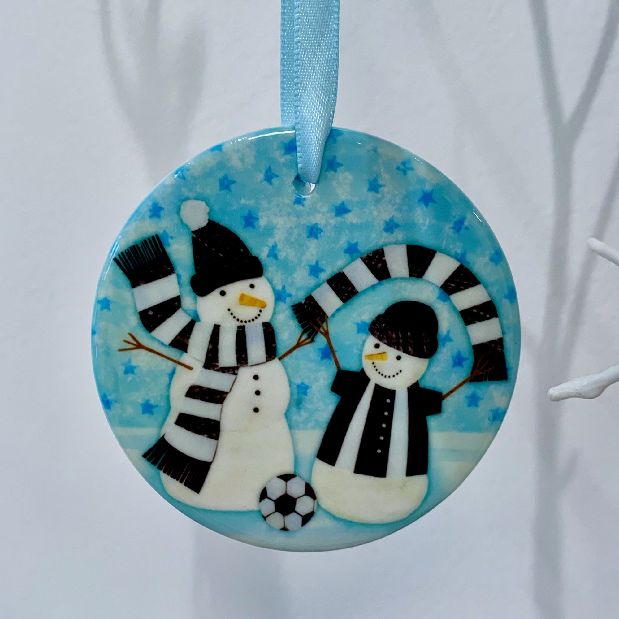 Circular ceramic decoration hung on a white branch. Happy snowmen wearing black and white scarves and football kit with a football at their feet. 