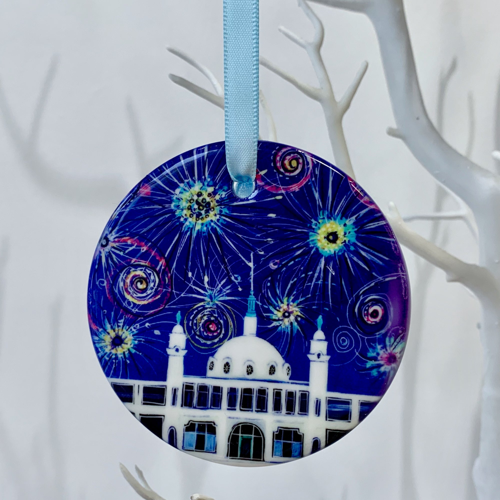 Circular ceramic decoration hung on a white branch. It features the Whitley Bay Spanish City Dome under a dark blue sky of colourful fireworks. 