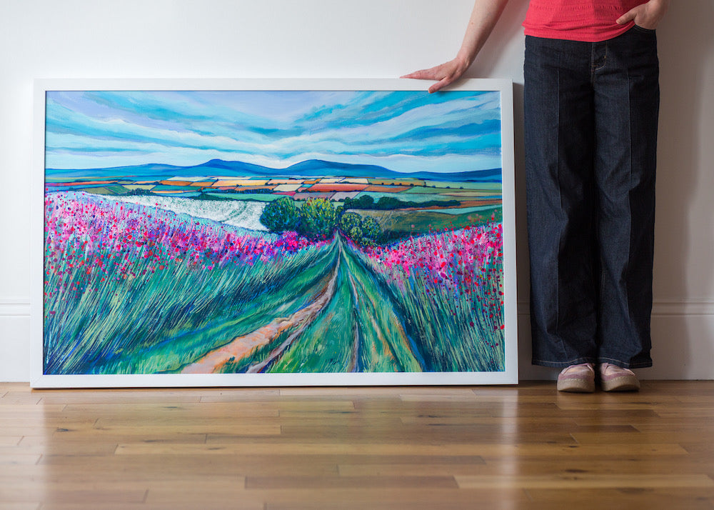 Artist stands beside panoramic painting featuring a path through pink wildflowers and a tapestry of fields below rolling blue hills on the horizon. The sky is a soft light blue with rippling clouds.