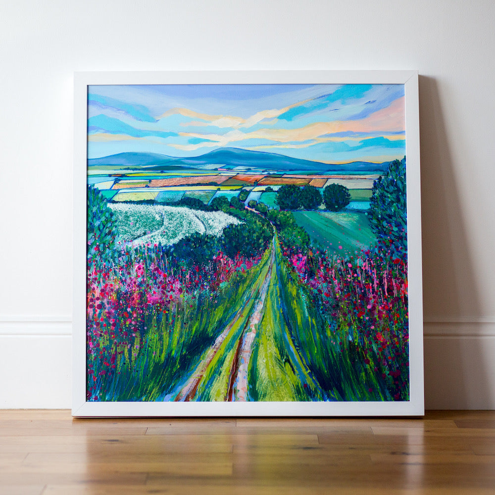 A narrow country path leads to the horizon through pink wildflowers and  green bushes on either side. A tapestry of fields lies just below the horizon and blue hills under a yellow and blue sky. 
