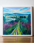 A narrow country path leads to the horizon through pink wildflowers and  green bushes on either side. A tapestry of fields lies just below the horizon and blue hills under a yellow and blue sky. 
