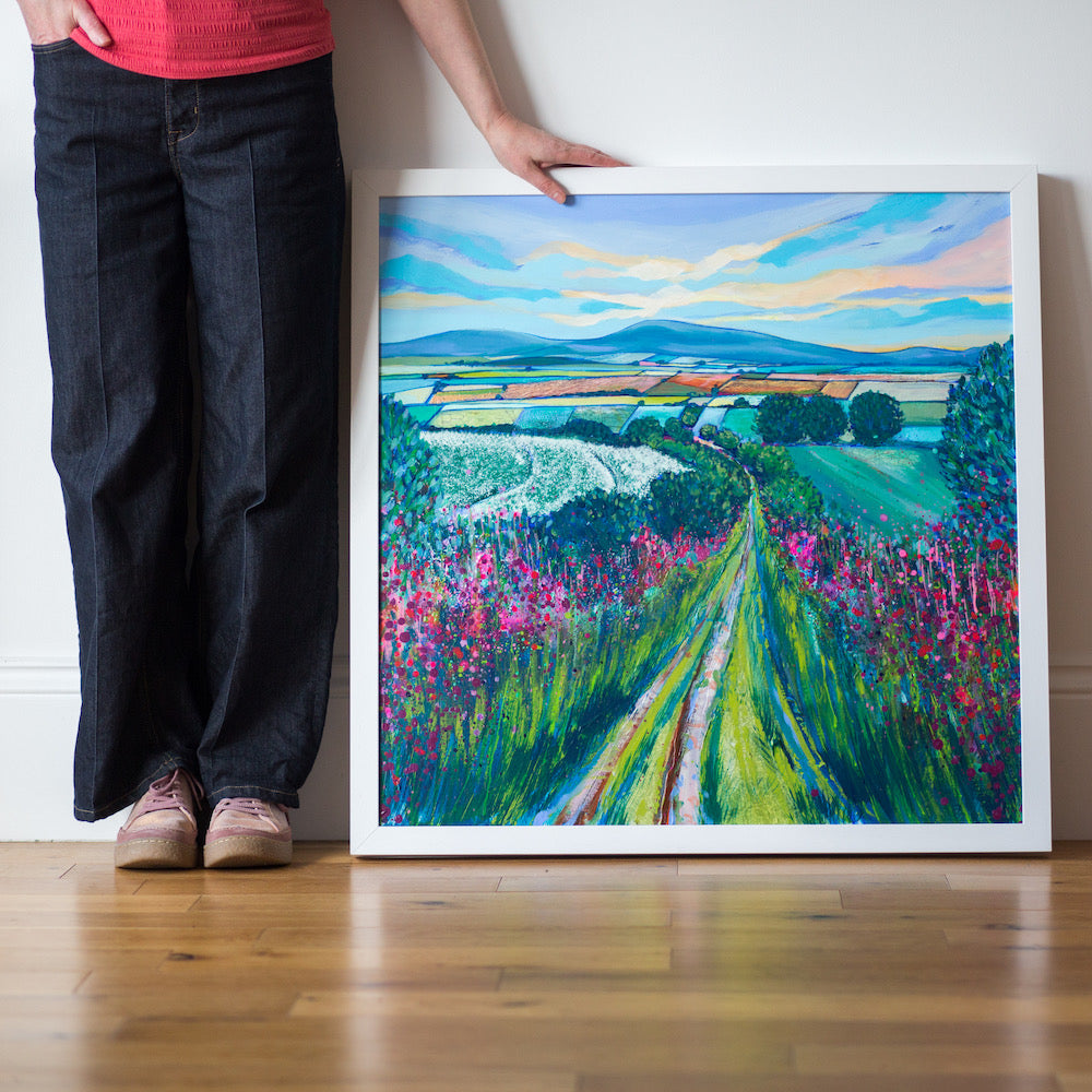 Artist's legs and hands alongside a painting featuring a narrow country path leads to the horizon through pink wildflowers and  green bushes on either side. A tapestry of fields lies just below the horizon and blue hills under a yellow and blue sky. 
