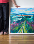 Artist's legs and hands alongside a painting featuring a narrow country path leads to the horizon through pink wildflowers and  green bushes on either side. A tapestry of fields lies just below the horizon and blue hills under a yellow and blue sky. 
