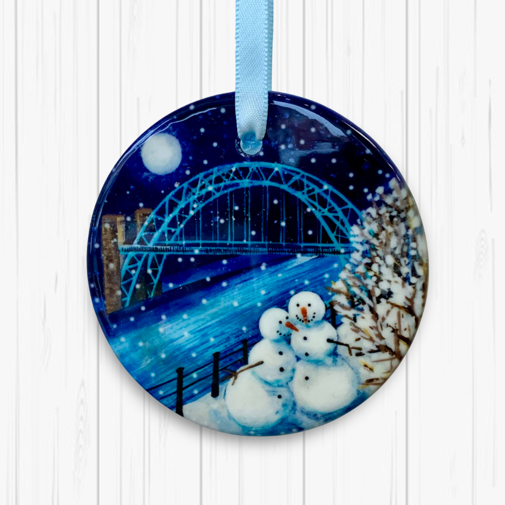 Circular ceramic hanging decoration on a white textured background. Two smiling snowmen stand side by side by the river with the Tyne Bridge in the background. The sky is dark with a full moon and snow is falling. 