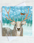 A Christmas Card featuring a stag and a robin perched on it's Antler. A green fir tree forrest is in the background with a flurry of snowfall overhead. 