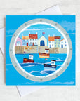 A coastal greetings card featuring a cute harbour with fishing boats on the sea.