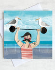 A greetings card of a dapper chap in a stripy vest lifting some weights on the sea front with two cheeky seagulls perched on top. 