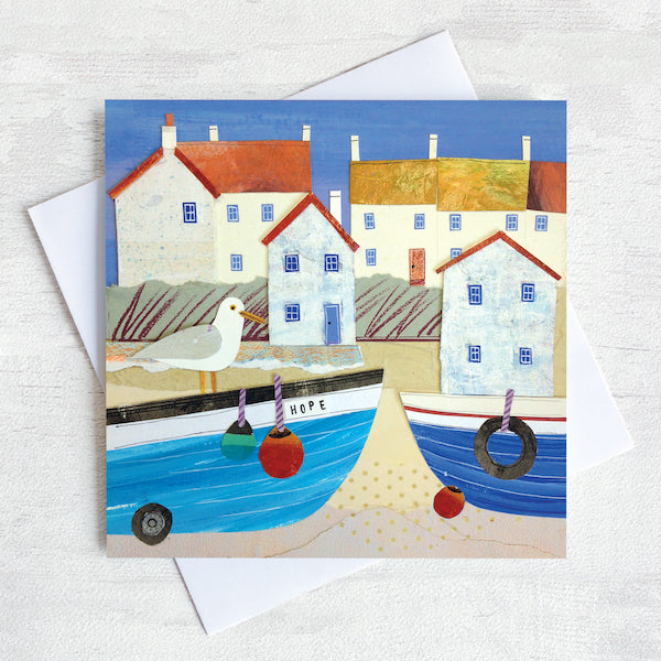 A greetings card featuring white harbour cottages with orange roofs and blue coble boats on the shore with a cheeky seagull perched. 
