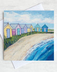 A greetings card featuring a painting of colourful beach huts on a sandy shore.