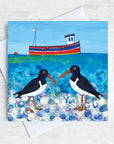 A charming greetings card from an original painting by Joanne Wishart, these quirky oystercatchers chat on a pebbled beach with a cute little fishing boat bobbing on a teal green sea.