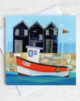 A greetings card featuring a bright red fishing boat in front of nostalgic net sheds on the harbour wall.