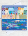 A quirky greetings card featuring a row of colourful cottages on a harbour wall with a shoal of colourful fish in the water.