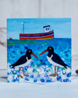 A charming greetings card featuring oystercatchers chatting on a pebbled beach with a cute little fishing boat bobbing on a teal green sea.